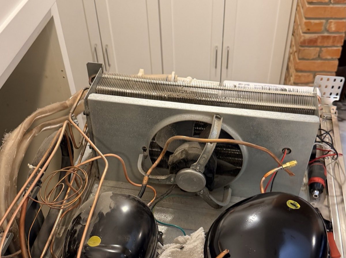 Machine compartment of a built-in refrigerator opened for service, showing both compressors, condenser coil, fan and copper line set