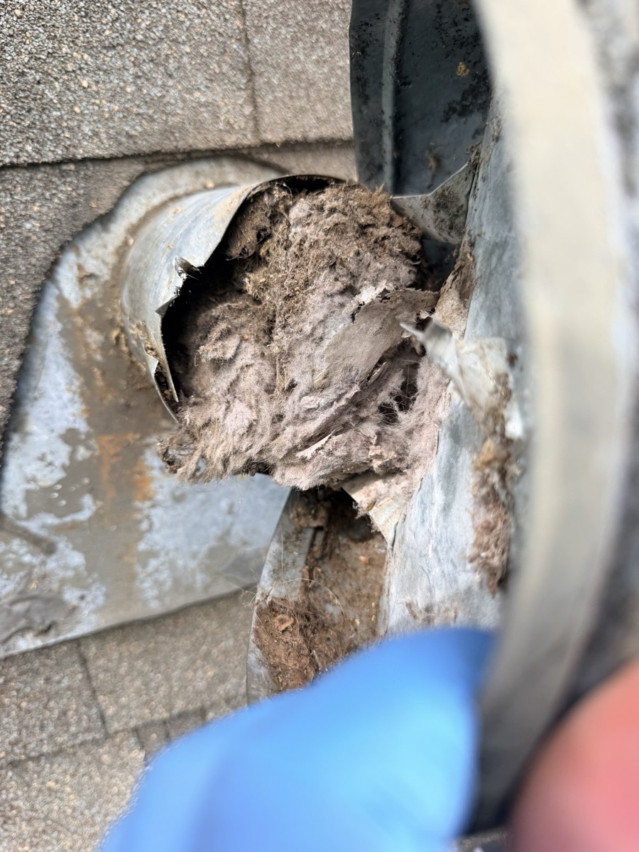 Dryer vent termination on a roof packed solid with lint, flap held back by a gloved hand