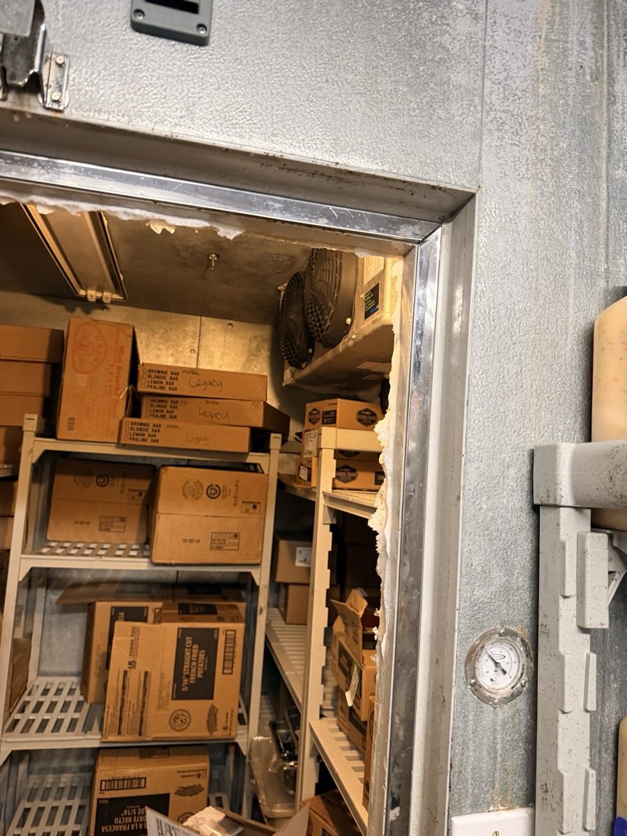 Interior of a stocked restaurant walk-in cooler seen through the doorway, with torn insulation on the door frame
